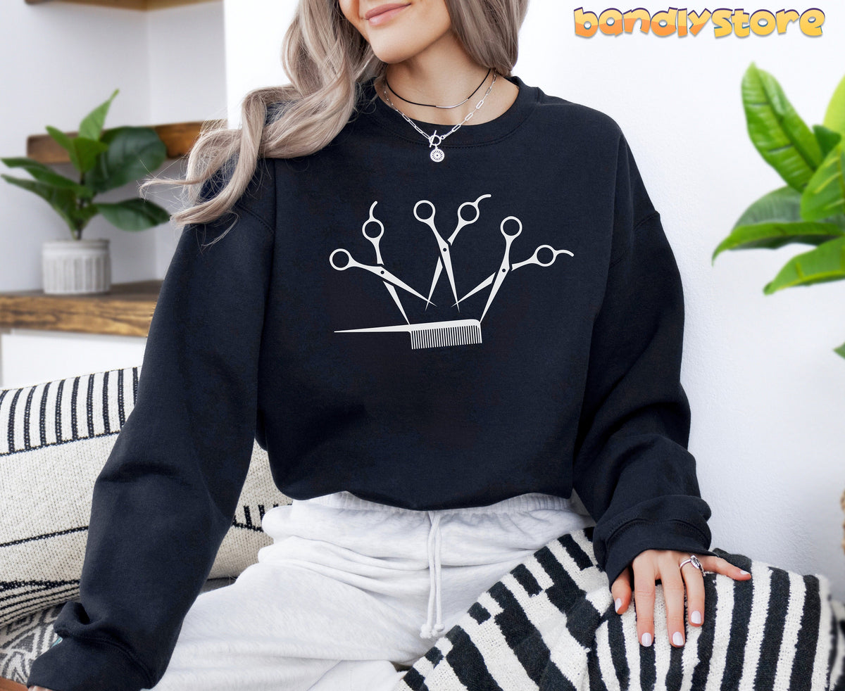 Hairstylist Crown Sweatshirt in black on a seated woman indoors beside plants and striped cushions.