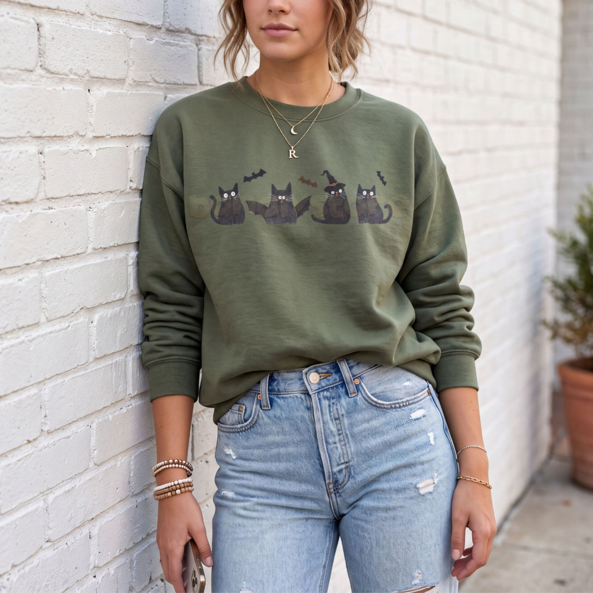 Halloween Black Cats And Bats Sweatshirt in military green on a person leaning against a white brick wall, front view.