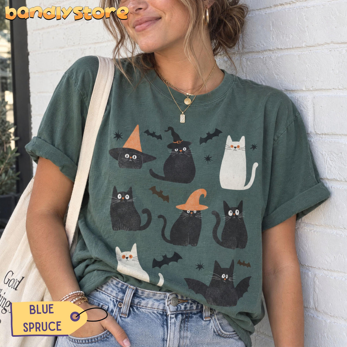 Cute Halloween Black Cats T-Shirt in Blue Spruce, worn by a woman against a white brick wall with tote bag and jeans.