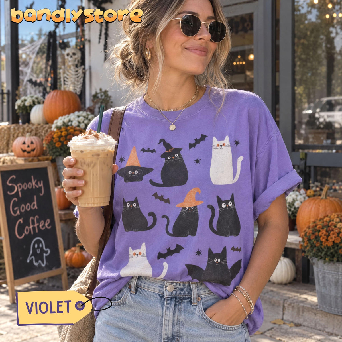 Cute Halloween Black Cats T-Shirt in violet on woman holding iced coffee outdoors among pumpkins and skeleton.