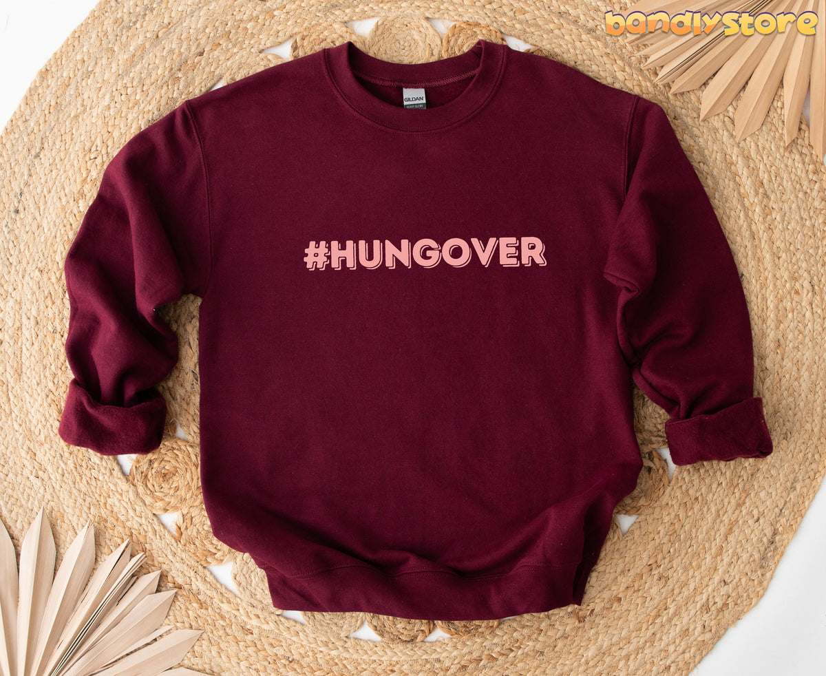 Hashtag Hangover Sweatshirt in maroon, flat lay on a round woven mat with dried palm leaves nearby.
