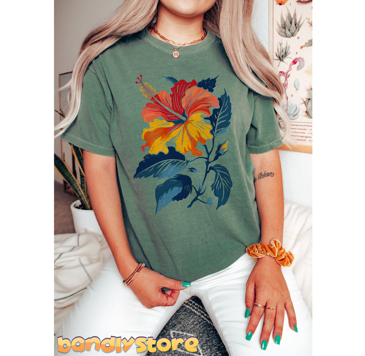 Hibiscus Flower T-Shirt in moss on a seated person indoors beside plants and wall posters.