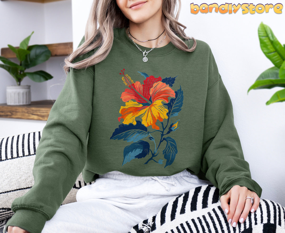 Hibiscus Flower Sweatshirt in military green, worn front-view indoors by a seated person near plants and shelves.