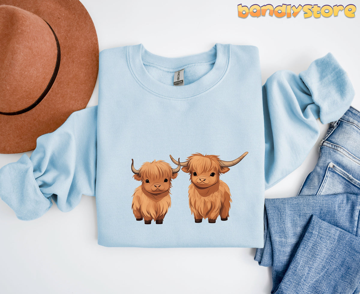 Highland Cow Sweatshirt in light blue laid flat beside a brown hat and jeans.