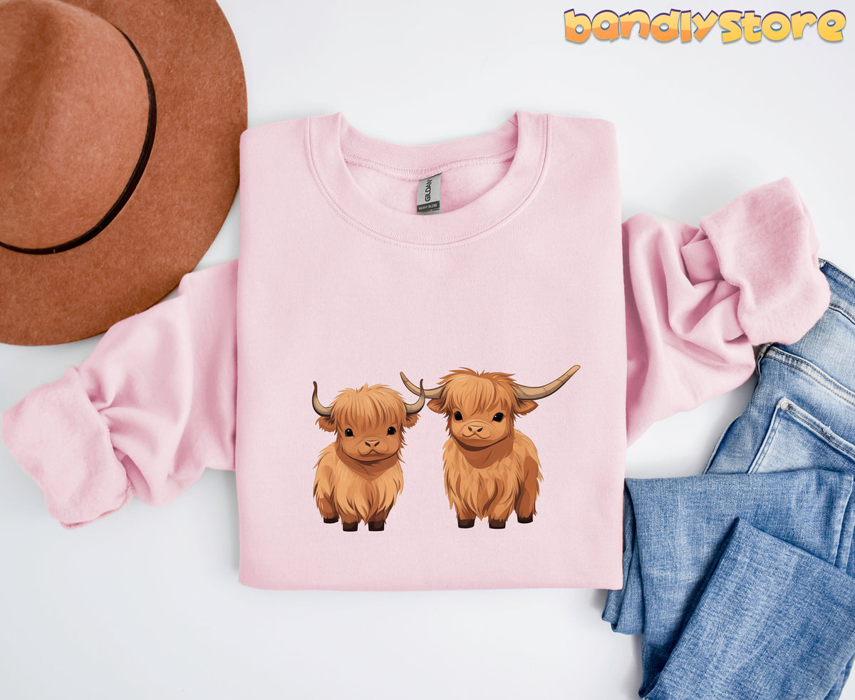 Highland Cow Sweatshirt in light pink laid flat beside a brown hat and blue jeans.