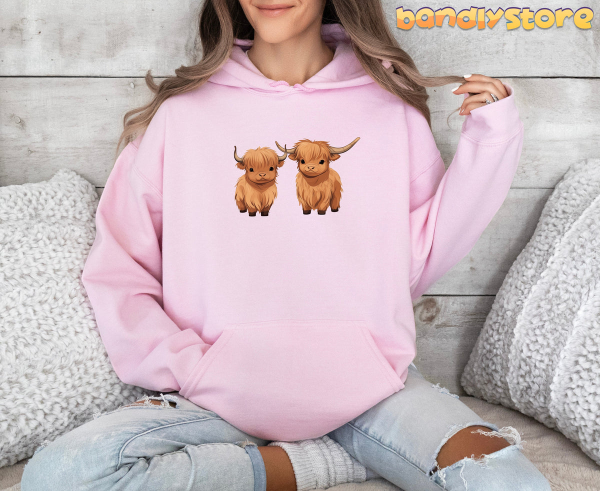 Highland Cow Hoodie in light pink, worn front-facing by a seated model amid white pillows on wood.