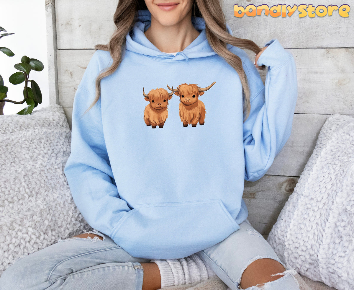 Highland Cow Hoodie in light blue, worn front-facing by a seated person amid pillows, plant and wood wall.