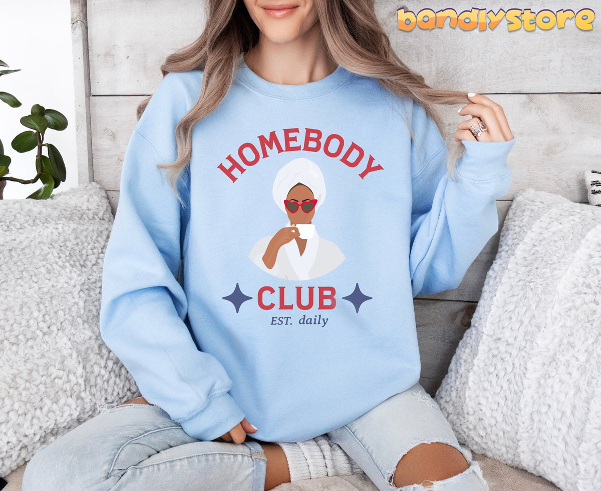 Homebody Club Sweatshirt in light blue on a seated person amid sofa pillows and a plant against a wood wall.