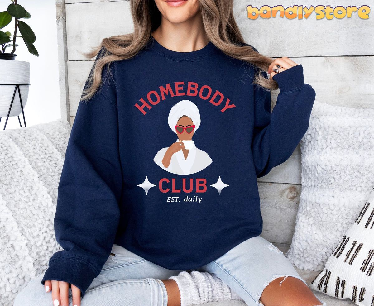 Homebody Club Sweatshirt in navy on a seated person by a white couch, pillows, plant and wood wall.