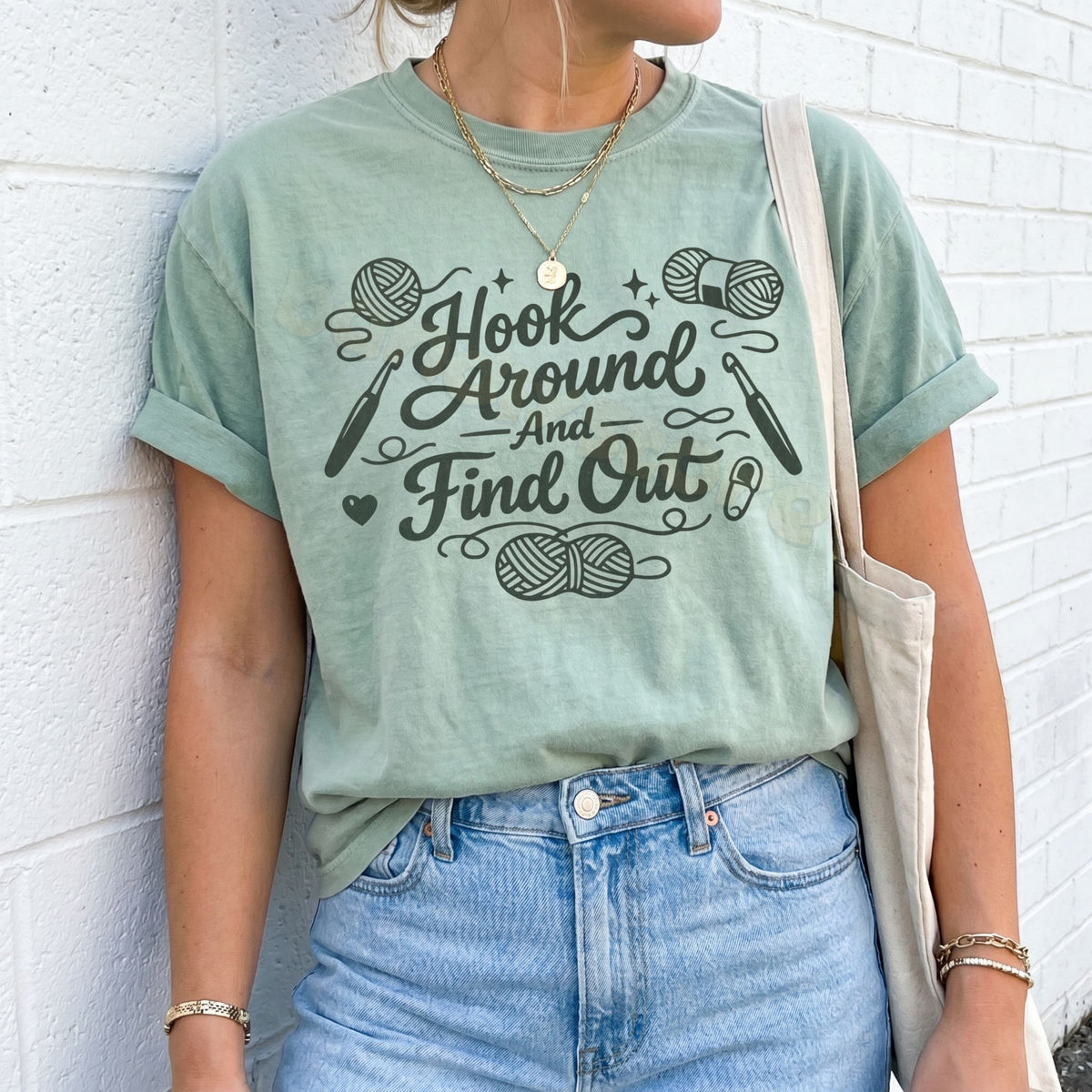 Hook Around And Find Out T-Shirt in bay on person against white brick wall with tote bag.