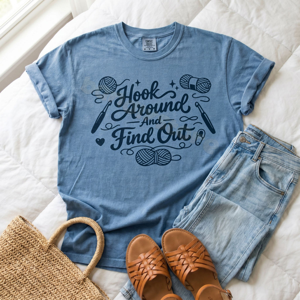 Hook Around And Find Out T-Shirt in Blue Jean flat-laid on white bedding with jeans, sandals, and a woven bag.