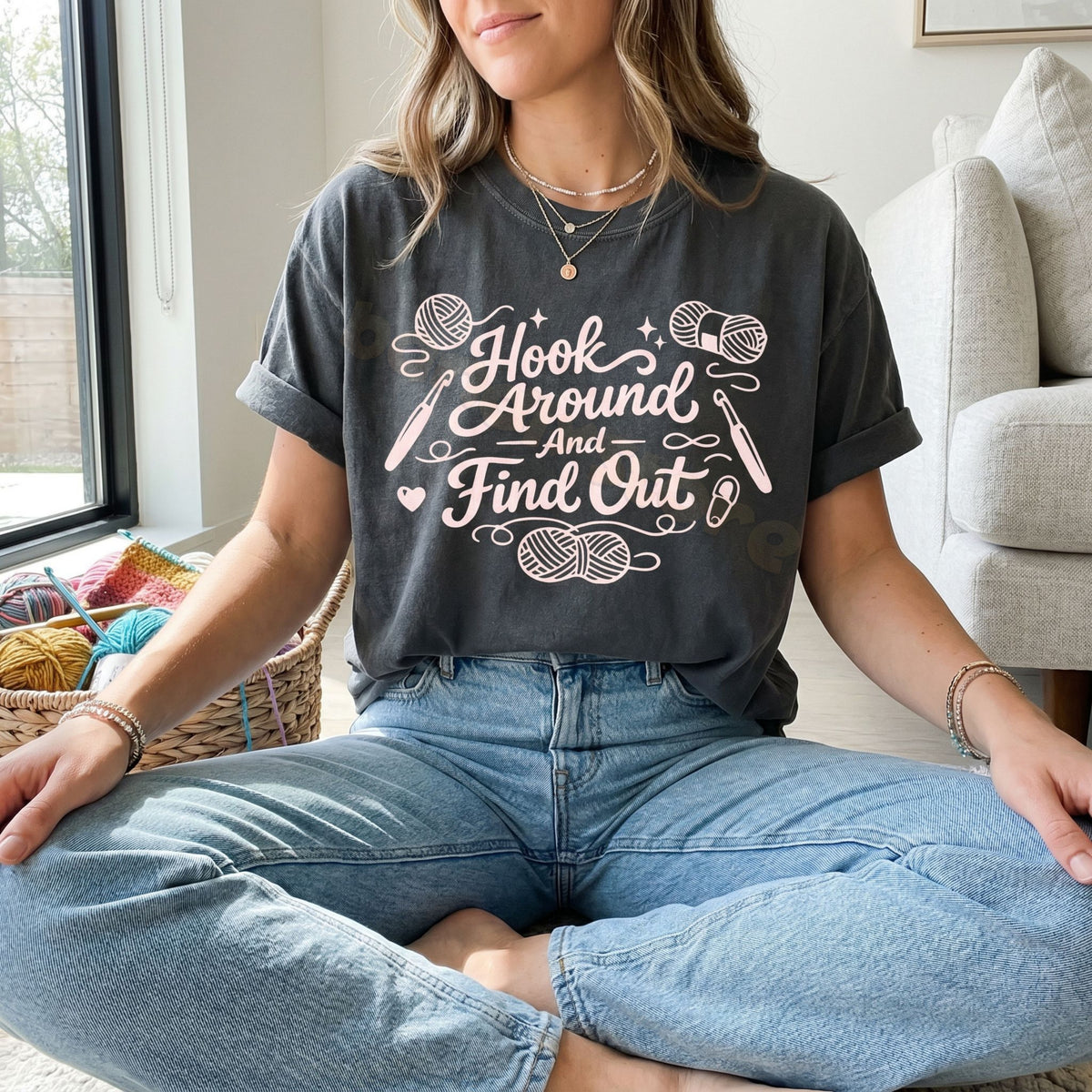 Hook Around And Find Out T-Shirt in Pepper on a woman sitting cross-legged indoors beside a yarn basket and sofa.
