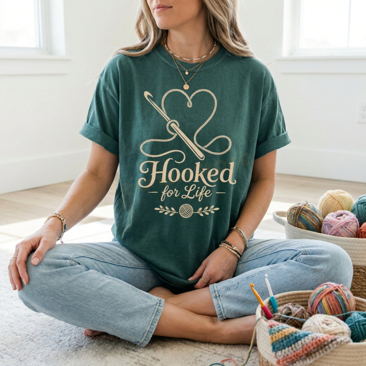 Hooked For Life T-Shirt in Blue Spruce on a woman sitting cross-legged amid yarn baskets.