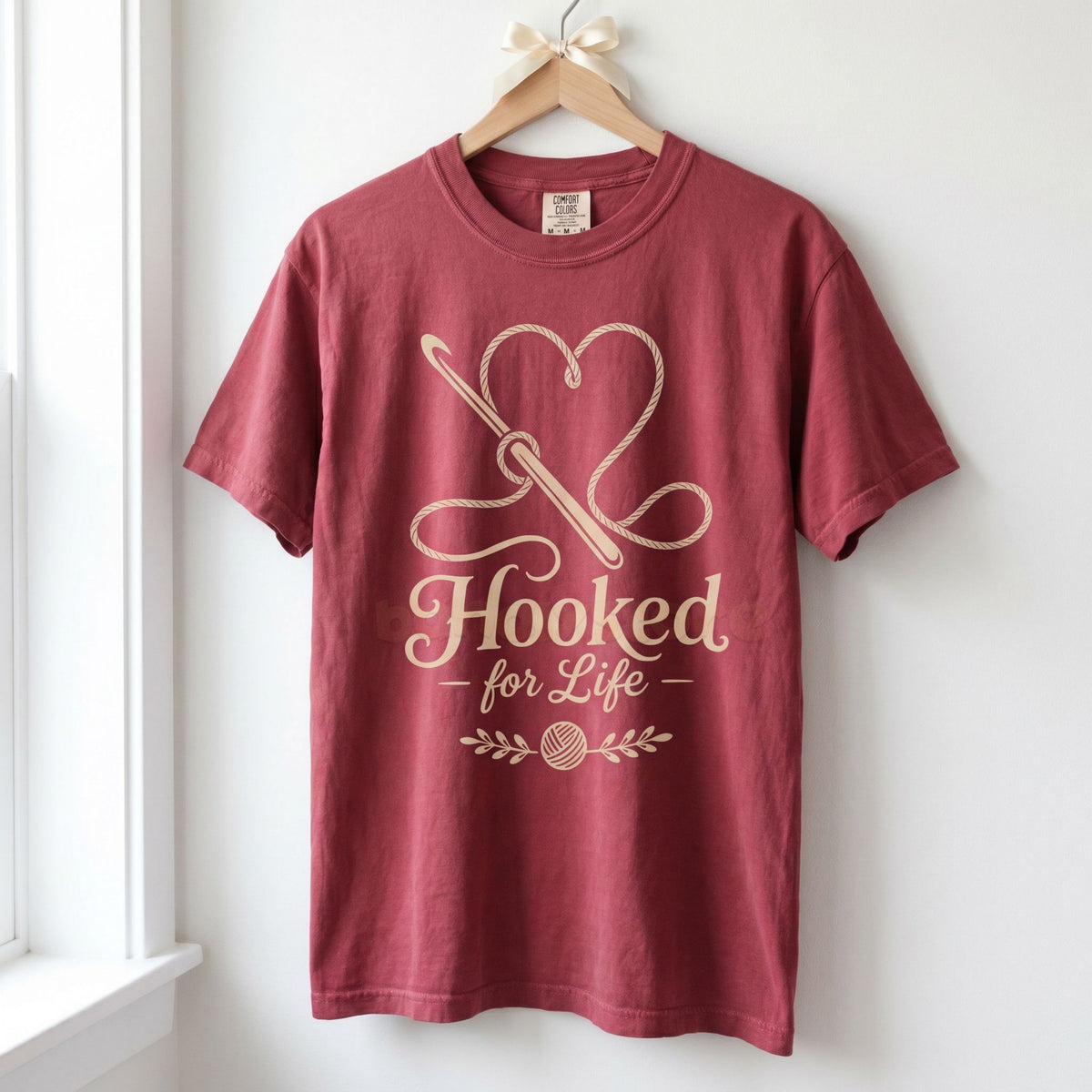 Hooked For Life T-Shirt in chili hanging front-facing on a wooden hanger against a white wall by a window.