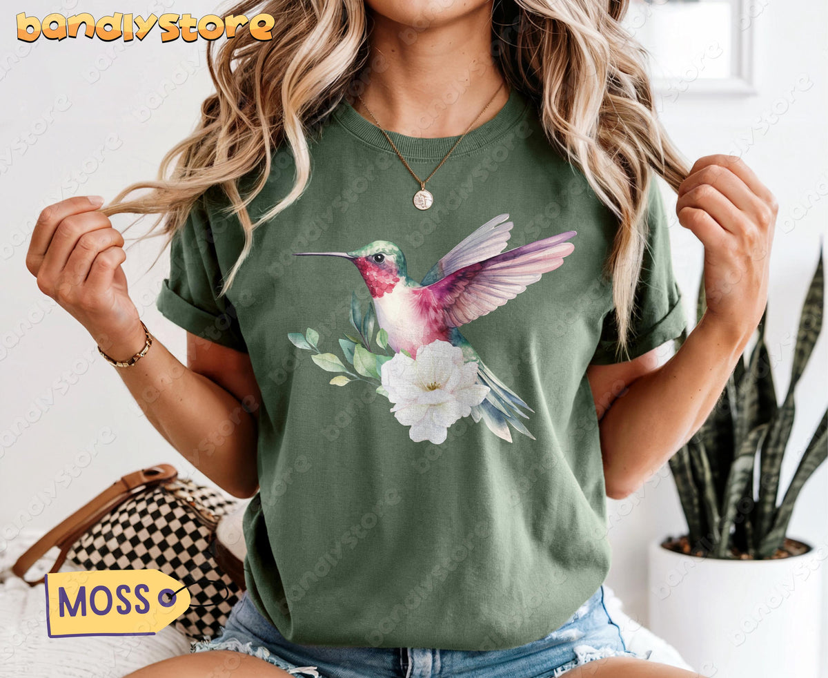 Hummingbird Flower T-Shirt in Moss, worn front-facing with a bag and plant visible nearby.
