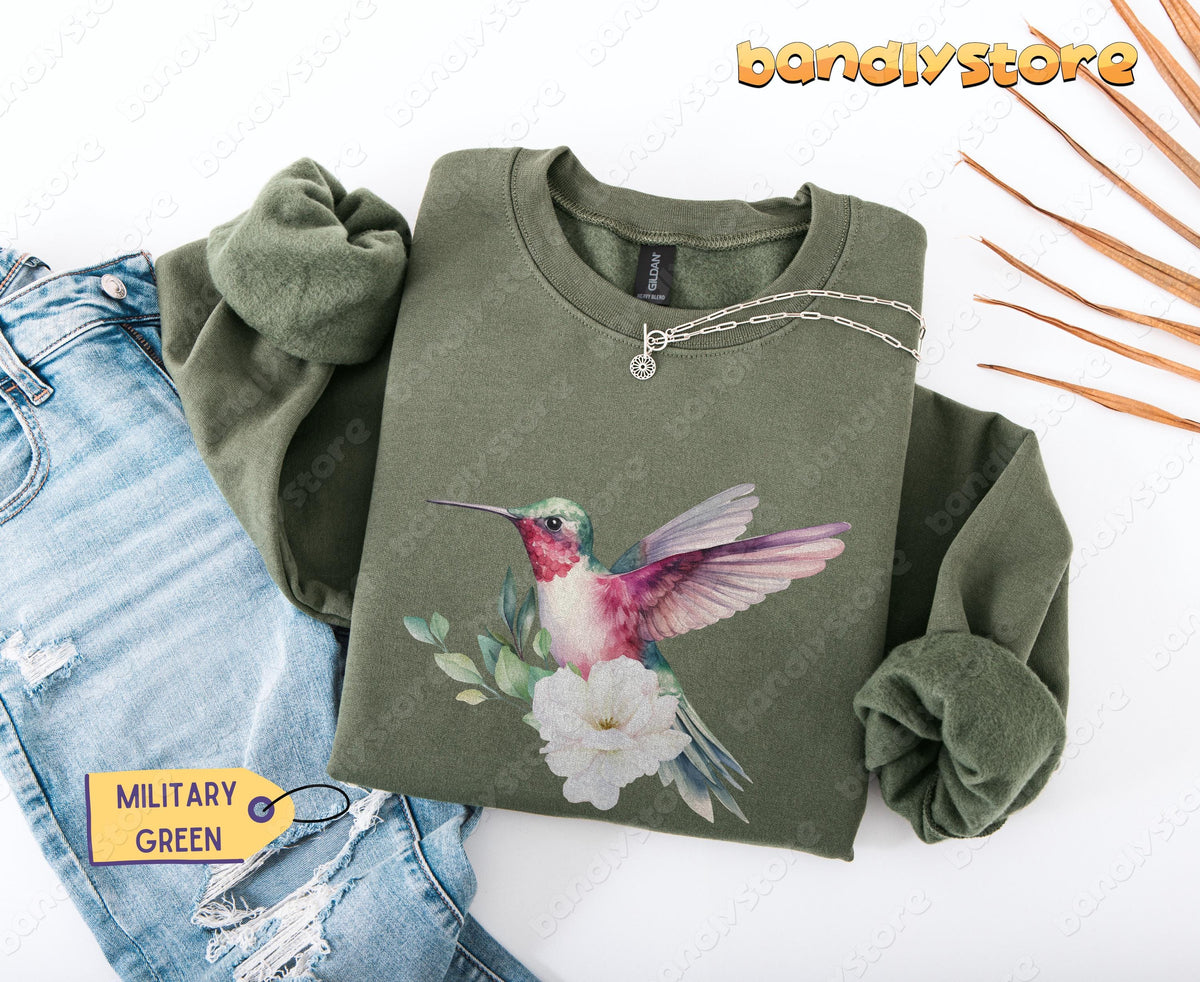 Hummingbird Flower Sweatshirt in military green flat-laid beside ripped jeans, silver necklace and dried leaves.
