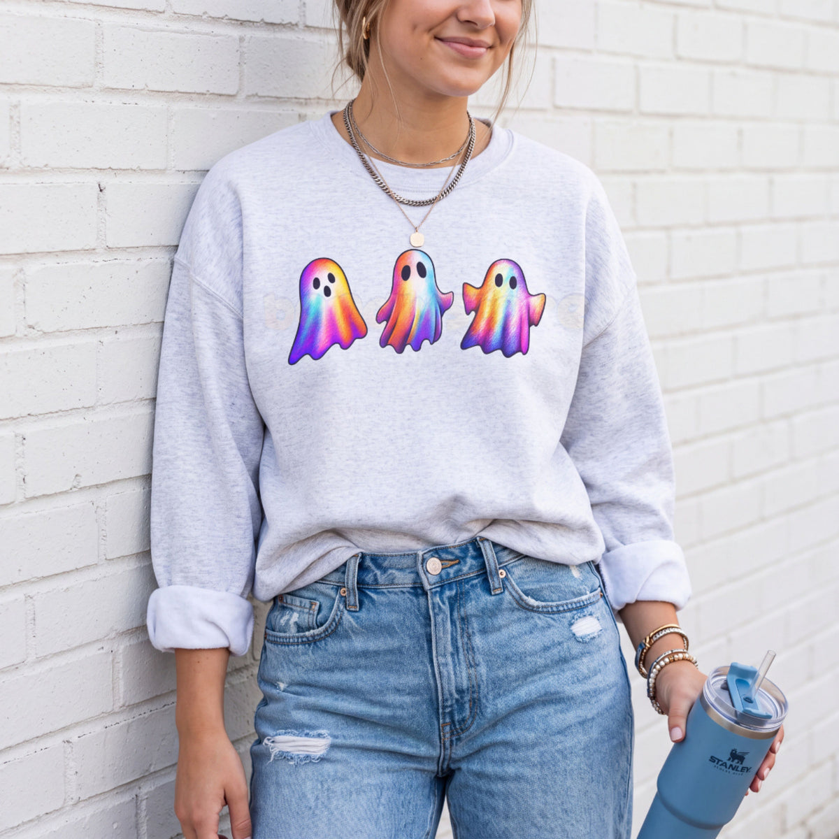 Iridescent Ghosts Sweatshirt in ash on a person leaning against a white brick wall holding a blue tumbler.