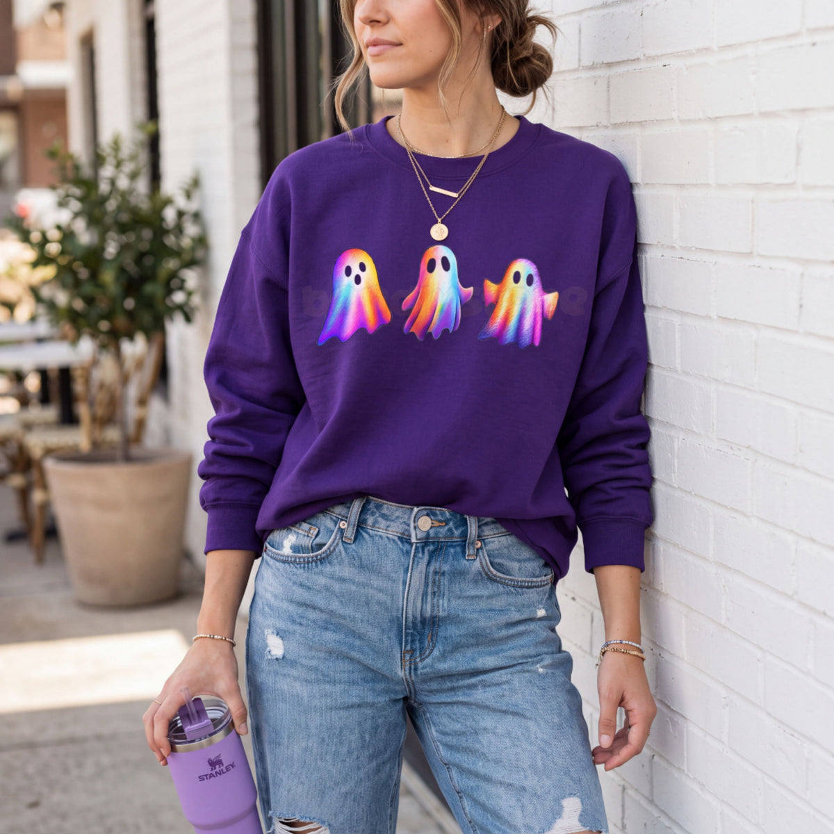 Iridescent Ghosts Sweatshirt in purple on a person leaning against a white brick wall outdoors holding a tumbler.