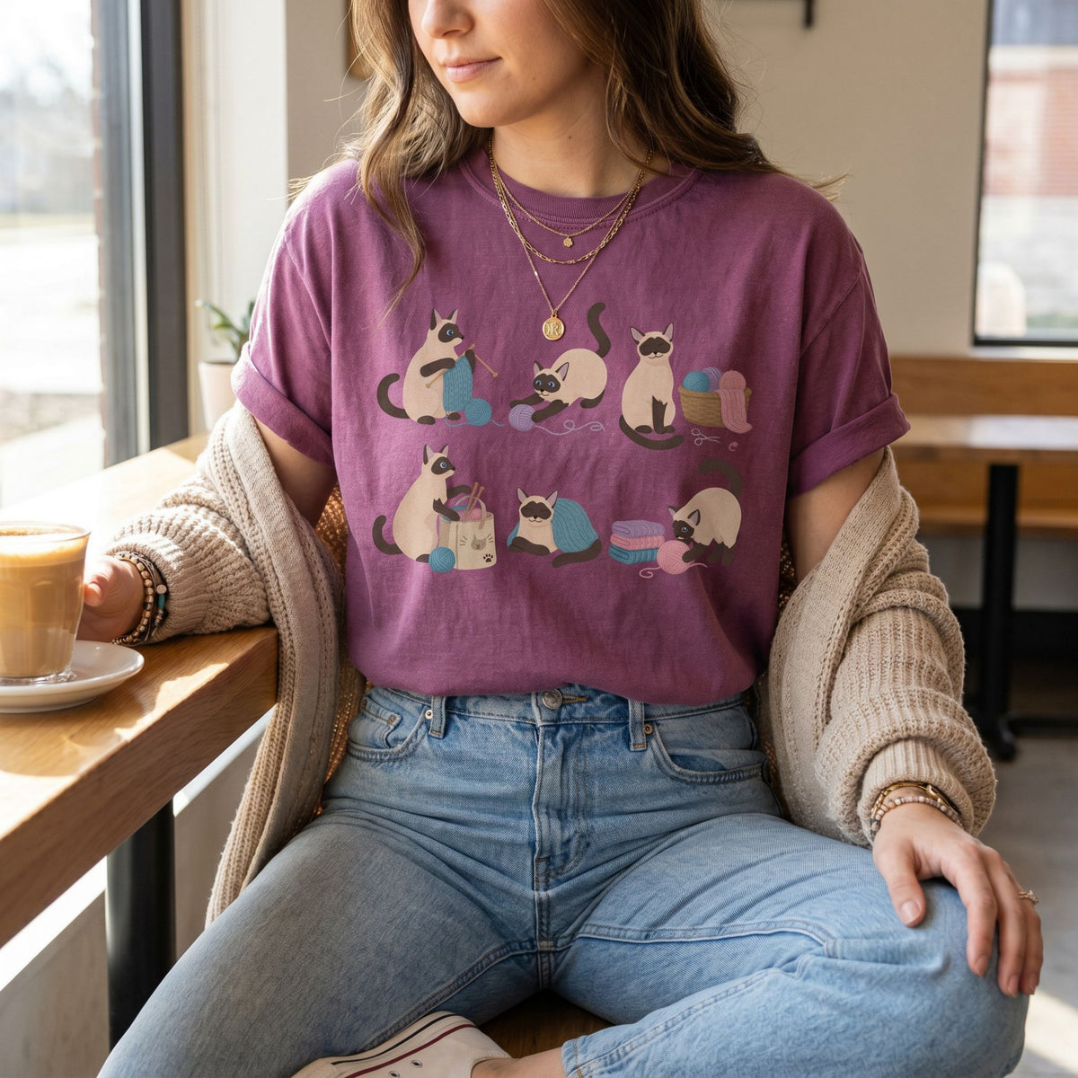 Funny Knitting Siamese Cats T-Shirt in berry worn by a woman seated at a wooden table with coffee by a window.