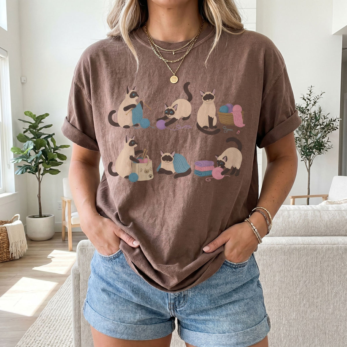 Funny Knitting Siamese Cats T-Shirt in espresso on a person standing frontally in a living room with plants and sofa.