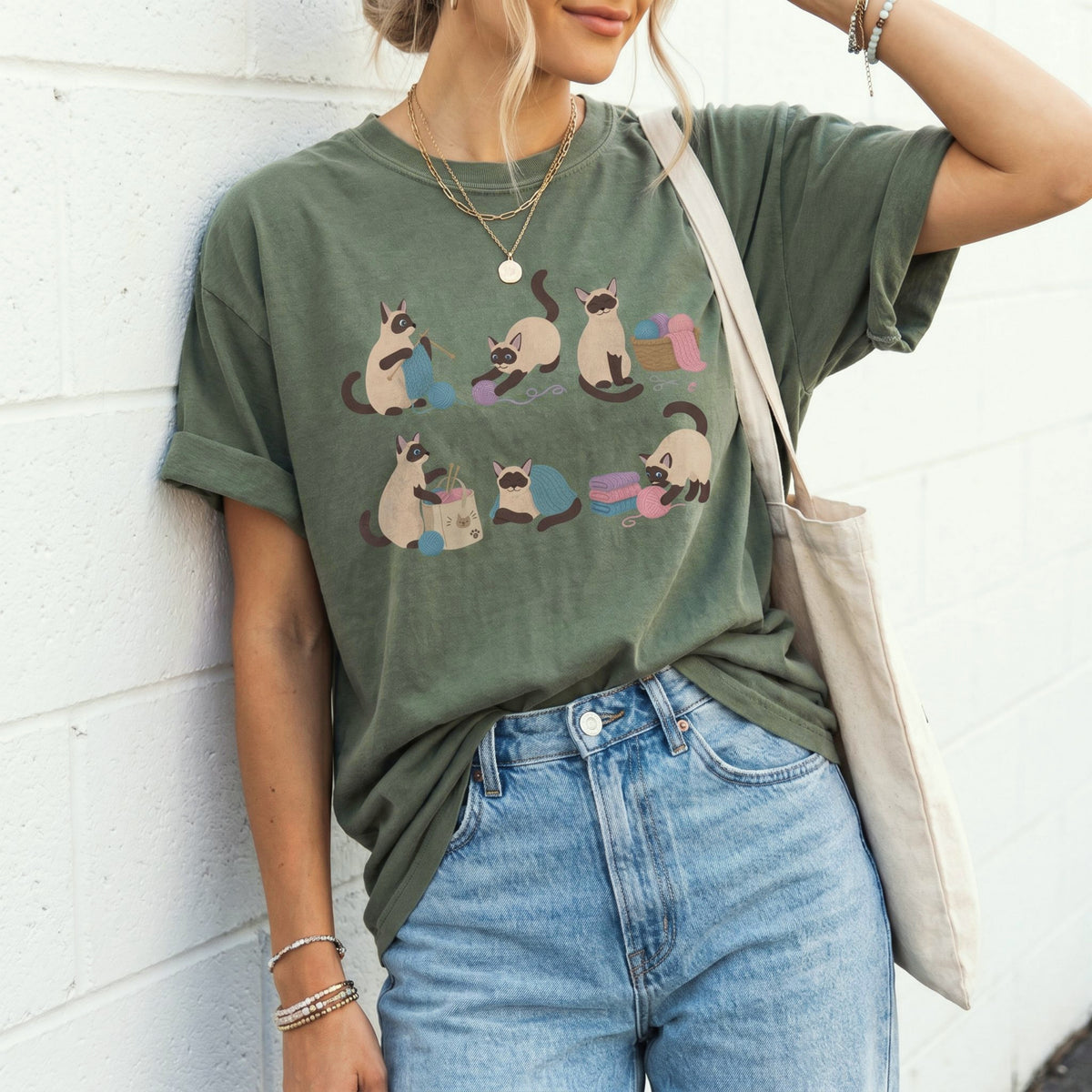 Funny Knitting Siamese Cats T-Shirt in moss on a person leaning against white brick wall with tote bag and jeans.