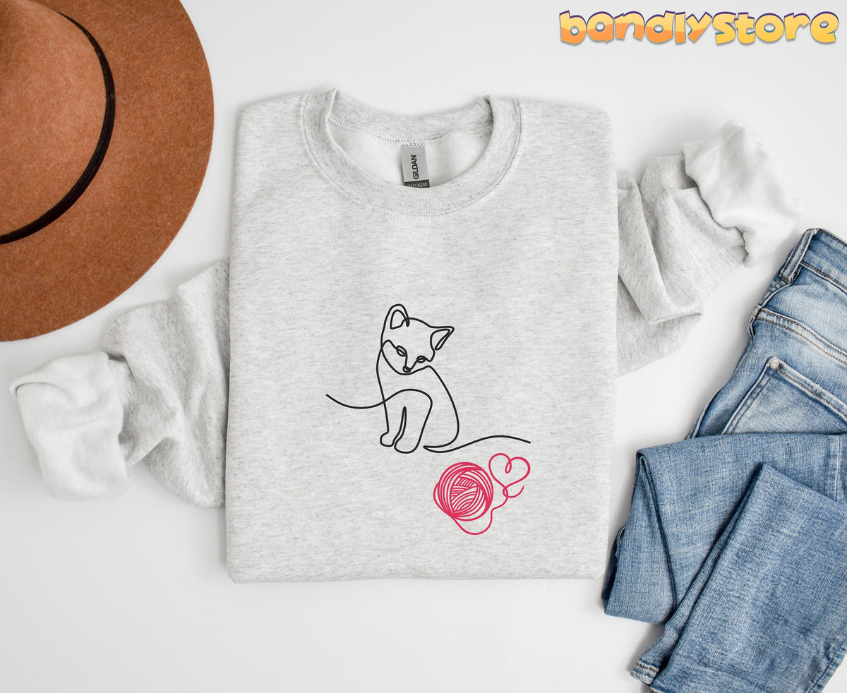Line Art Kitten Sweatshirt in ash, folded flat from above beside a brown hat and blue jeans.