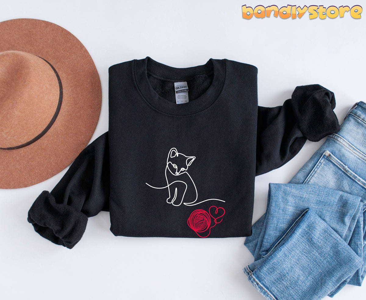 Line Art Kitten Sweatshirt black crewneck flat-lay with brown hat left and blue jeans right.