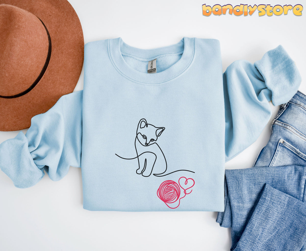 Line Art Kitten Sweatshirt in light blue folded flat top-down beside brown hat and blue jeans.