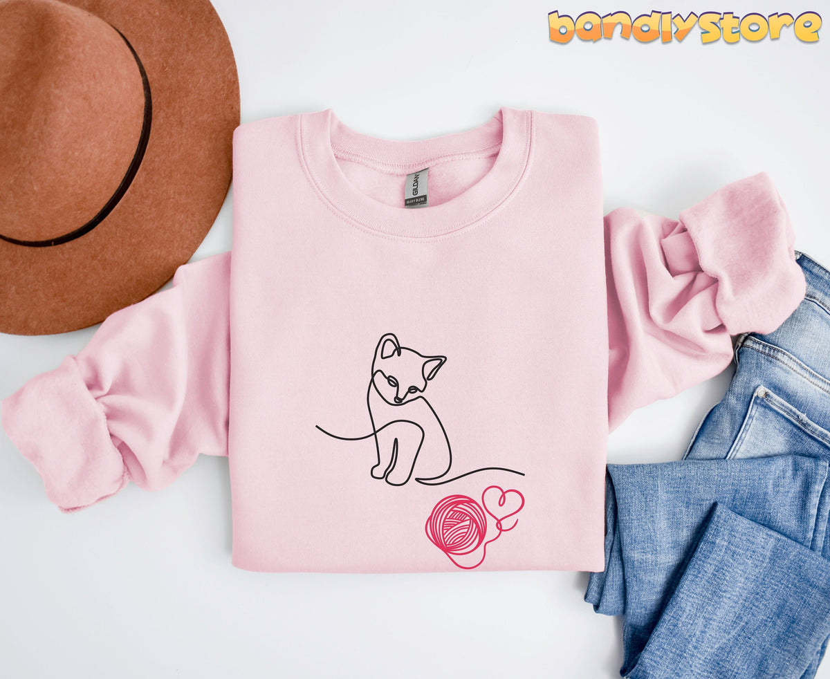 Line Art Kitten Sweatshirt in light pink laid flat from above beside a brown hat and blue jeans.