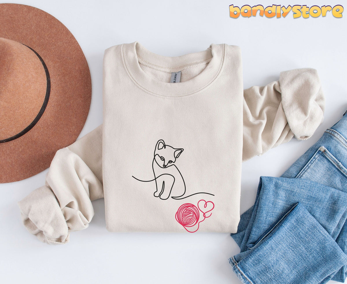 Line Art Kitten Sweatshirt in sand folded flat beside a brown hat and blue jeans on white.