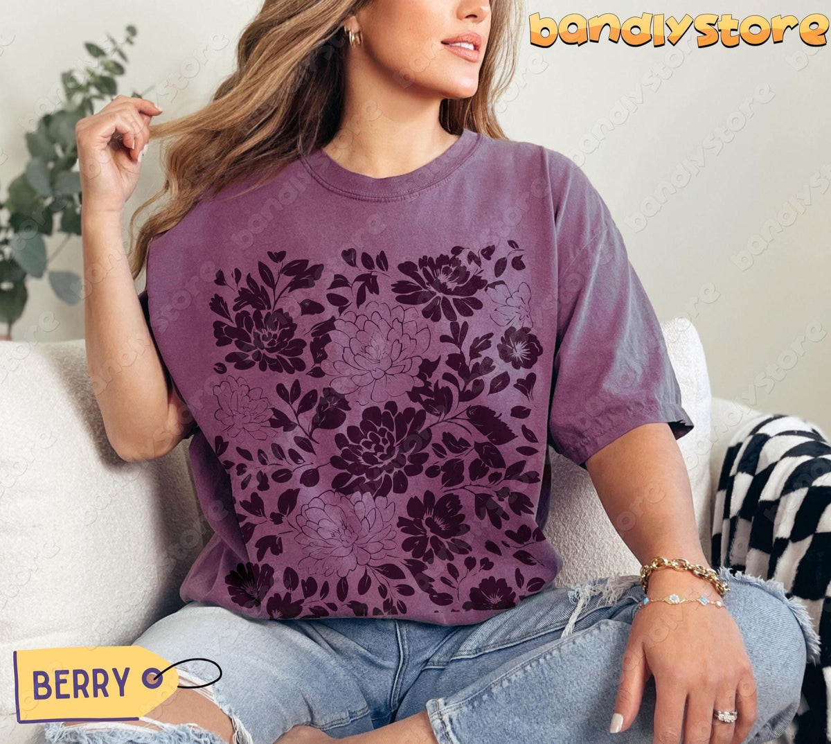Linocut Flowers T-Shirt in berry, modelled seated on a light sofa with plants and a checked throw nearby.