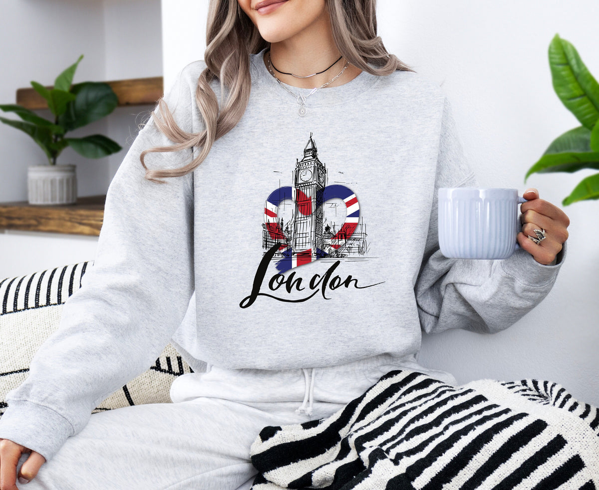 London Big Ben Sweatshirt in ash, worn by a seated woman holding a mug amid plants and striped textiles.