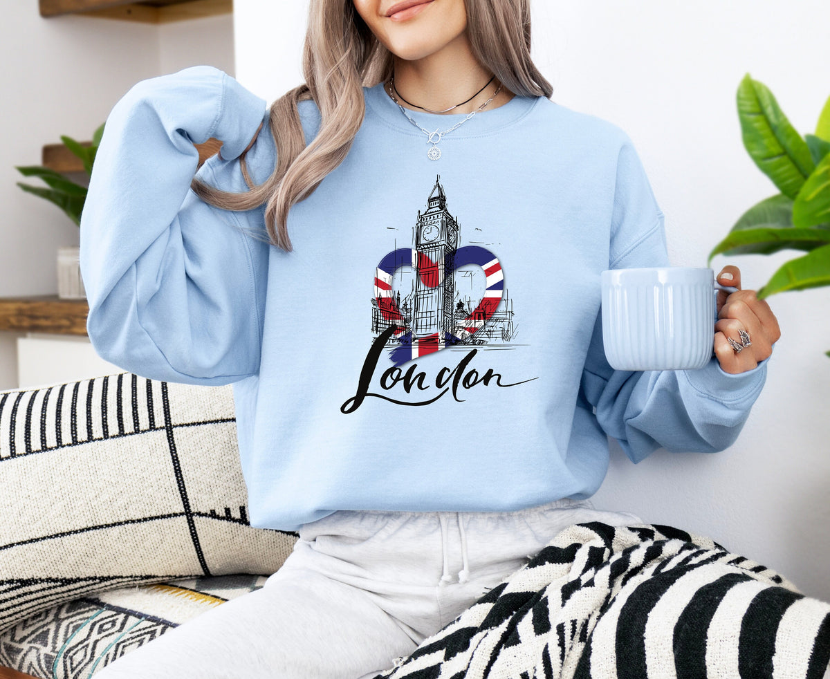 London Big Ben Sweatshirt in light blue worn by a seated woman holding a mug amid cushions and plants.