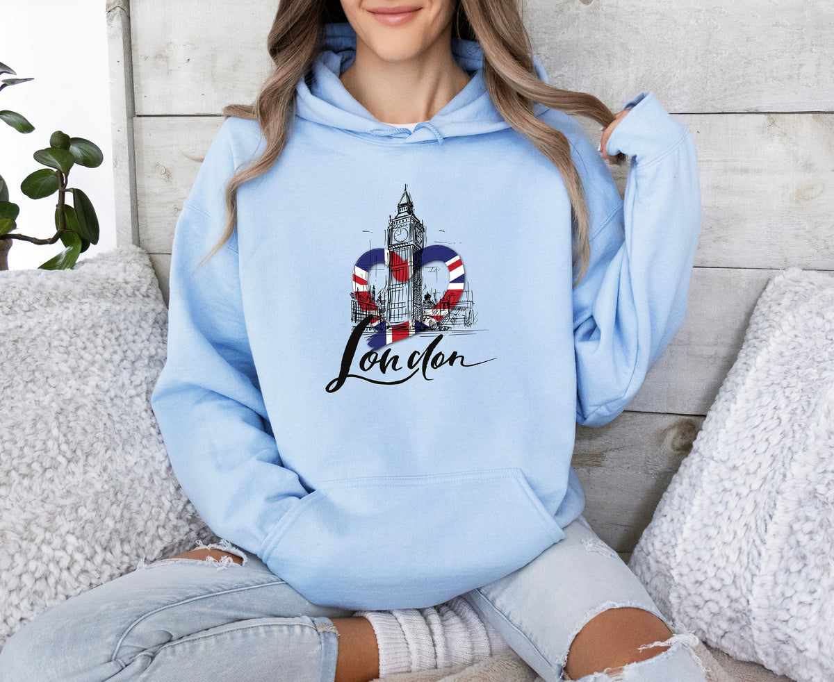London Big Ben Hoodie in light blue, worn by a seated person amid pillows and a plant against a wood wall.