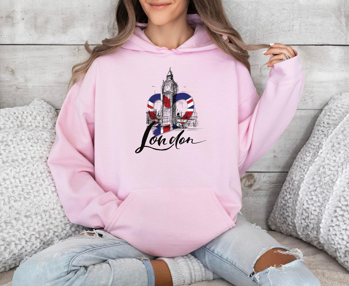 London Big Ben Hoodie in light pink worn by a seated person against a wooden wall with white pillows.