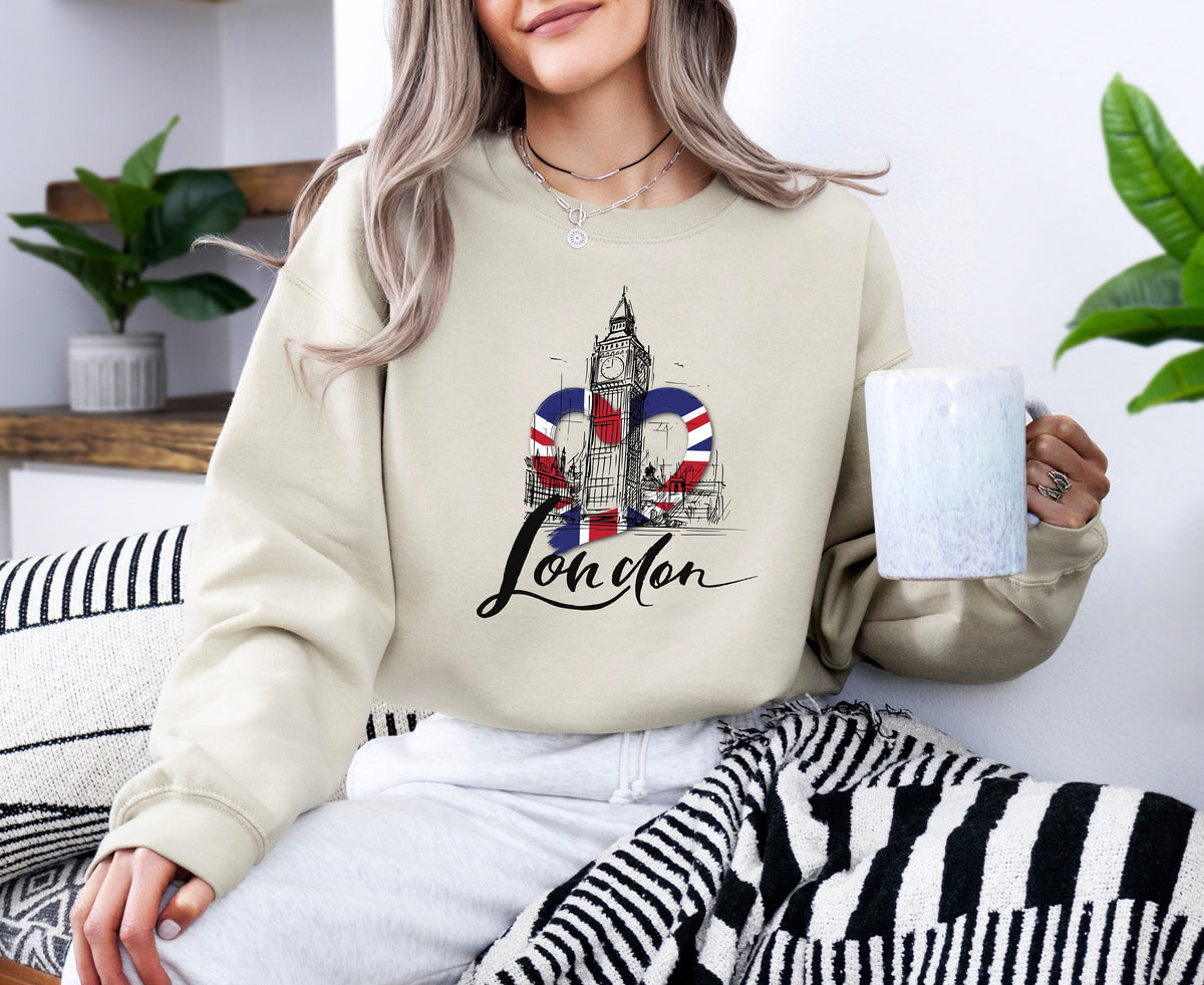 London Big Ben Sweatshirt in sand, worn by a seated woman holding a mug amid plants and striped textiles.