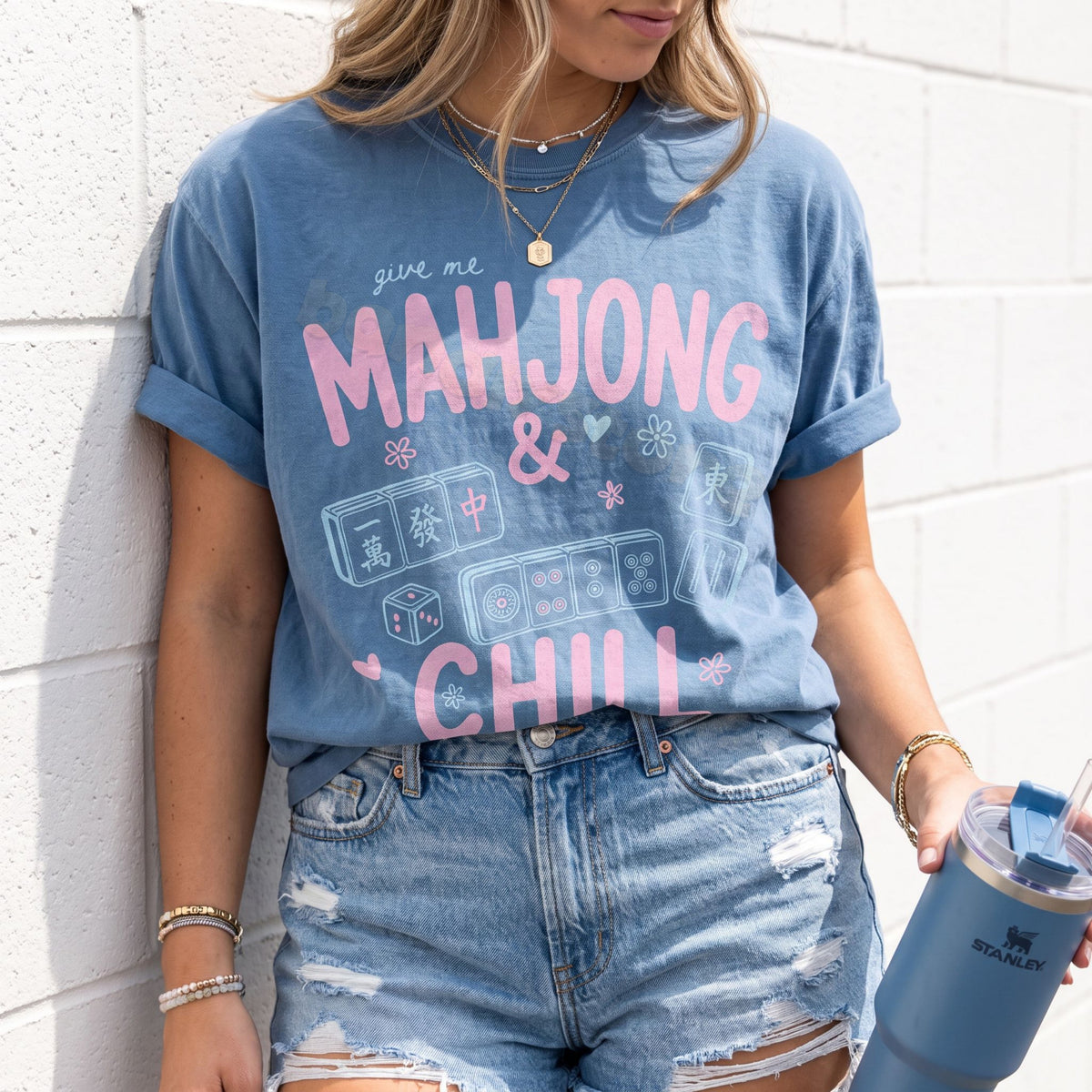 Mahjong And Chill T-Shirt in Blue Jean, worn by a person leaning on a white brick wall holding a blue tumbler.