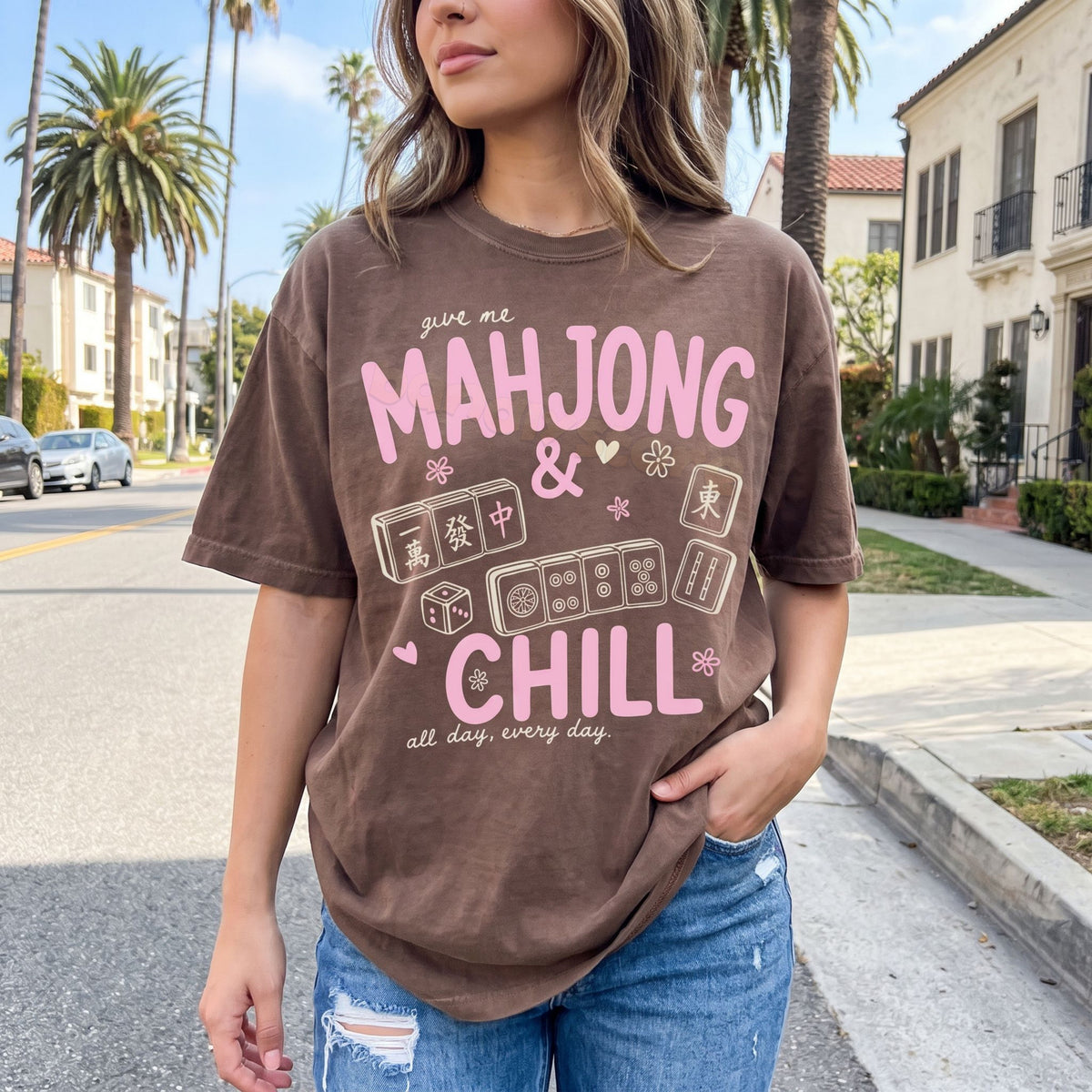 Mahjong And Chill T-Shirt in espresso shown front-on on a woman standing on a street with palm trees and houses.