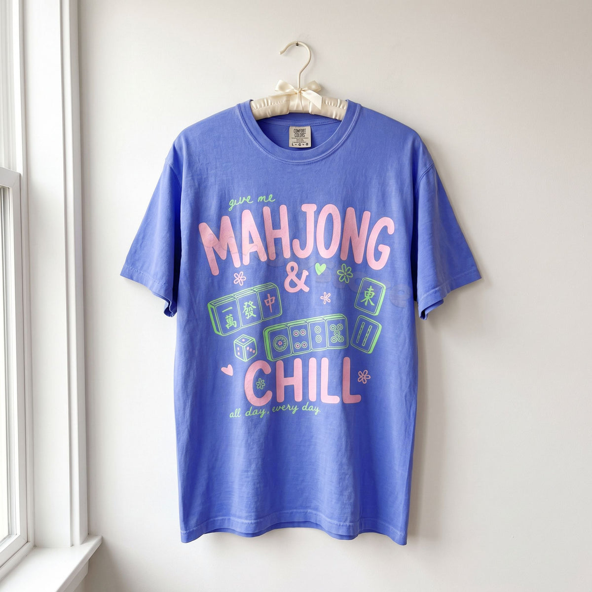 Mahjong And Chill T-Shirt in Flo Blue hanging on a hanger against a white wall beside a window.