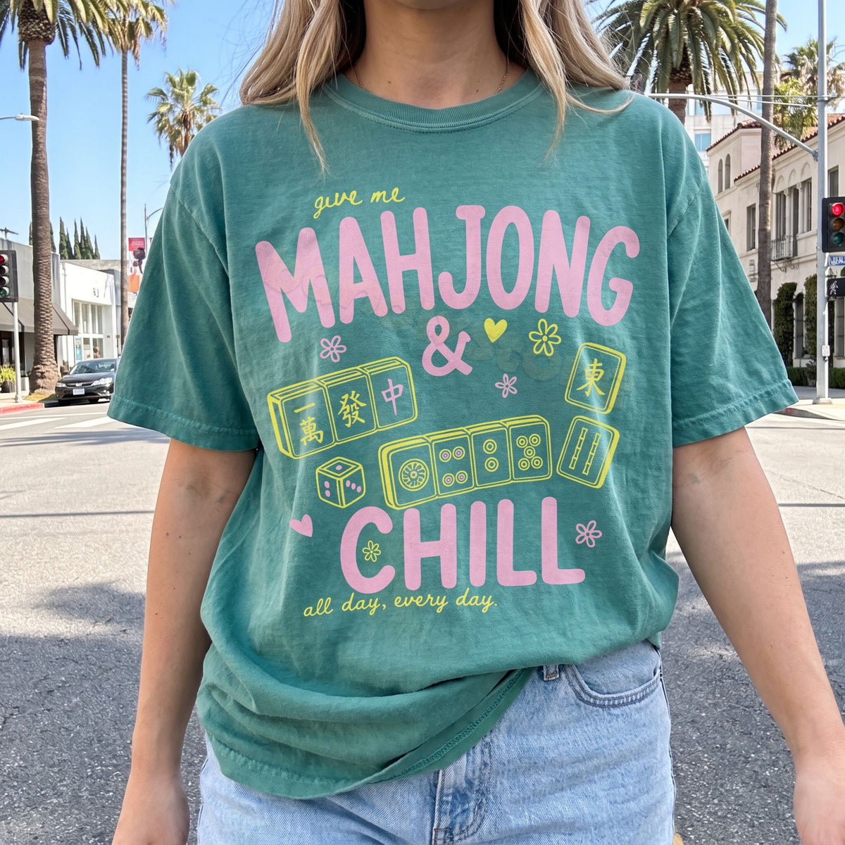 Mahjong And Chill T-Shirt light green on a person standing outdoors on a city street with palm trees, front view.