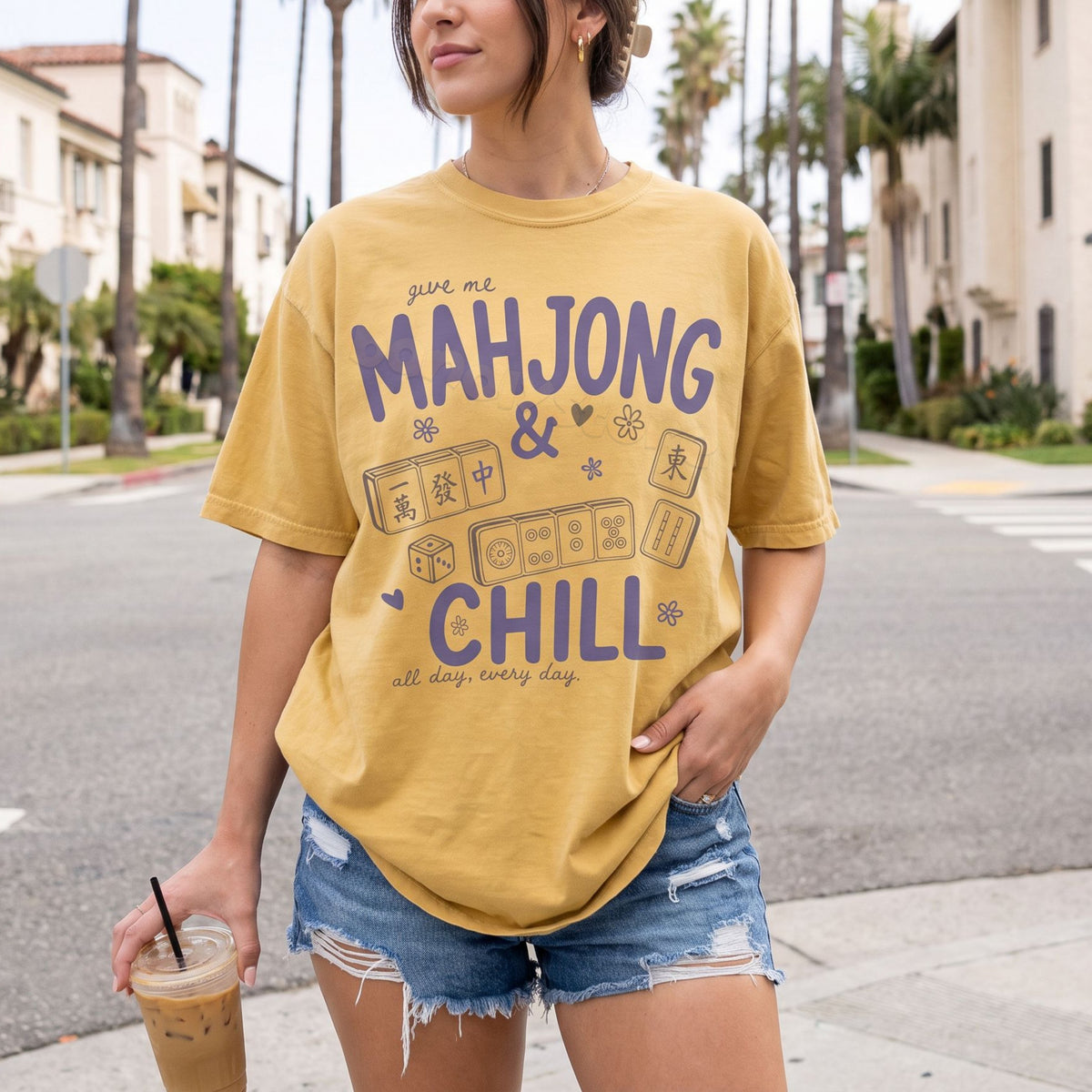 Mahjong And Chill T-Shirt in mustard on a person facing forward on a palm-lined street holding an iced coffee.