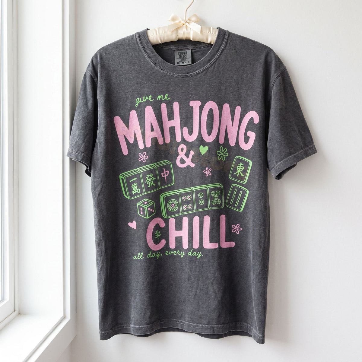 Mahjong And Chill T-Shirt in Pepper hanging on a hanger against a white wall by a window.