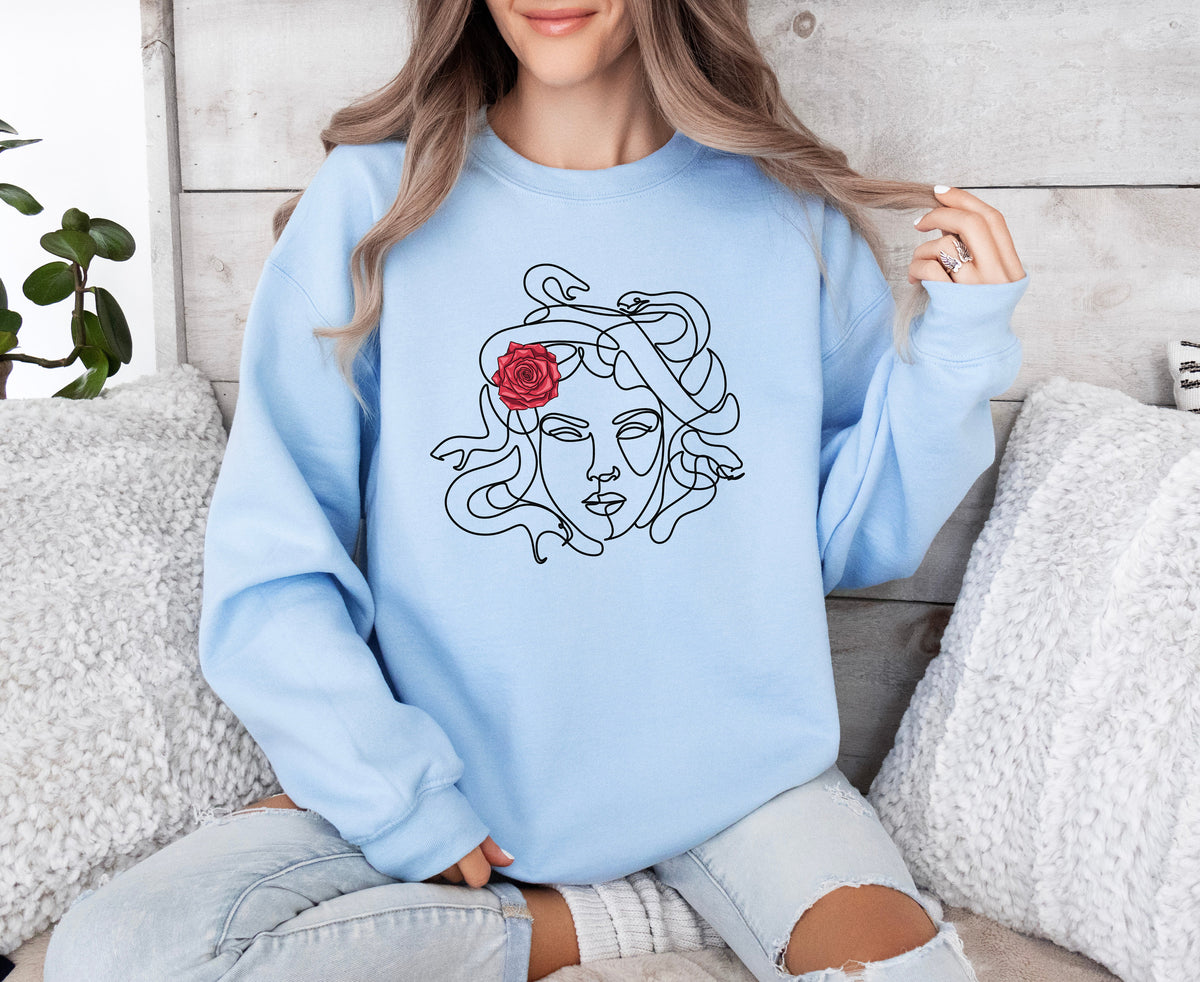 Medusa Sweatshirt in light blue, worn front-facing by a seated person amid pillows, plant and wood wall.