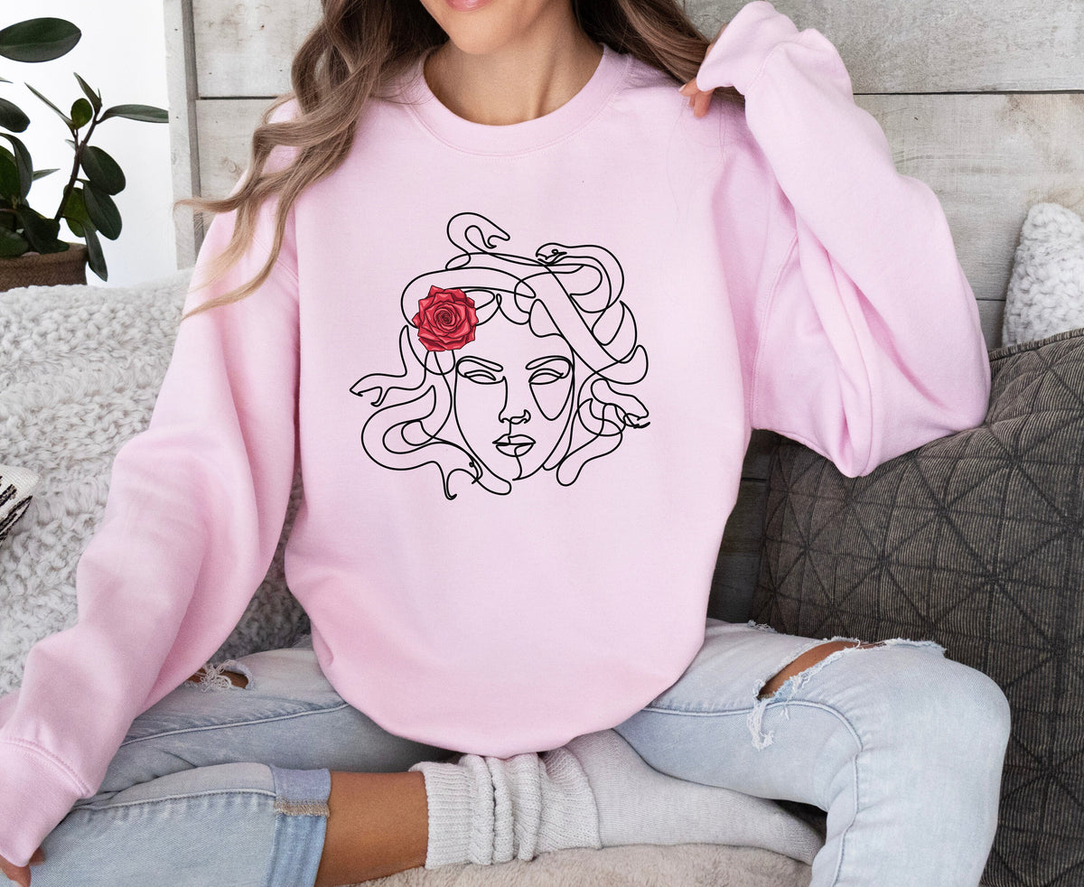 Medusa Sweatshirt in light pink worn frontally by a seated person beside pillows and a plant on wood paneling.