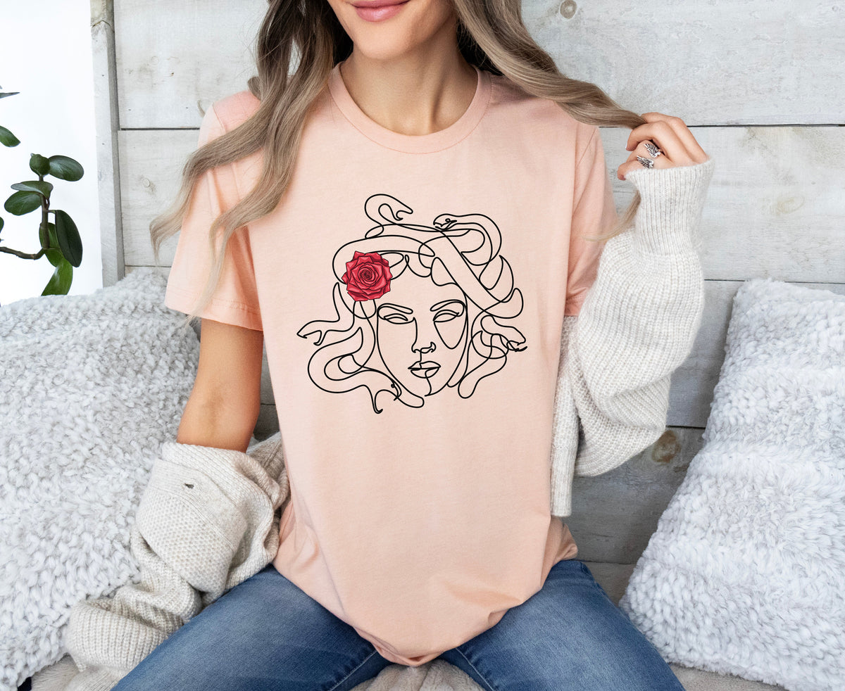 Medusa Head T-Shirt in heather peach on a seated woman against wood wall with fluffy pillows and plant.