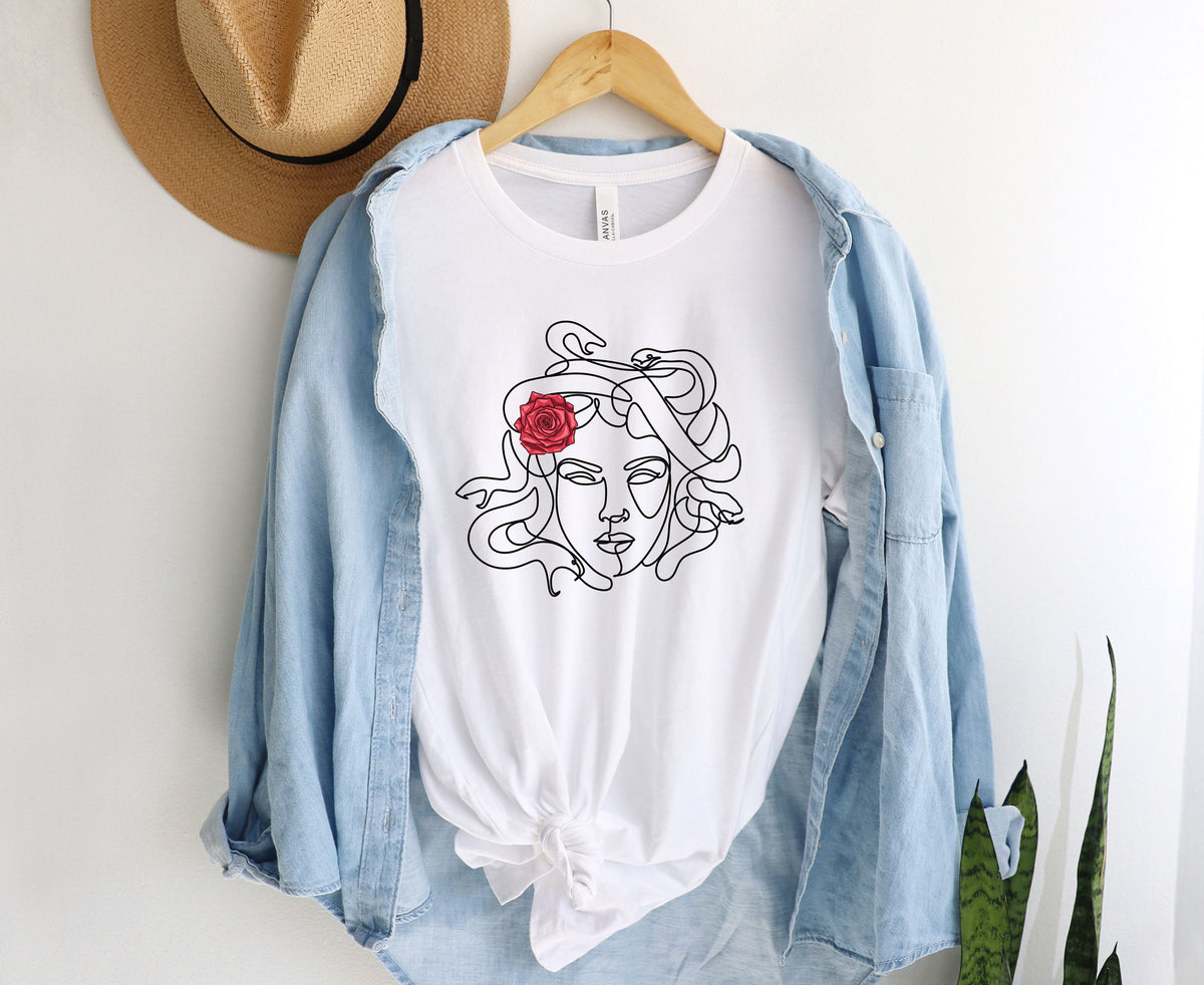 Medusa Head T-Shirt in white hung on a wooden hanger against a wall with straw hat, draped denim shirt and plant leaves.