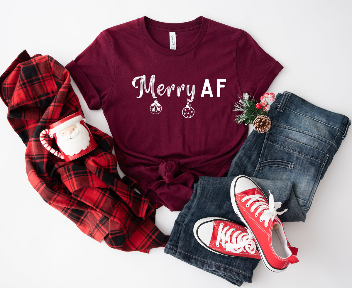Merry AF T-Shirt in maroon laid flat with plaid shirt, Santa mug, jeans, sneakers and greenery.