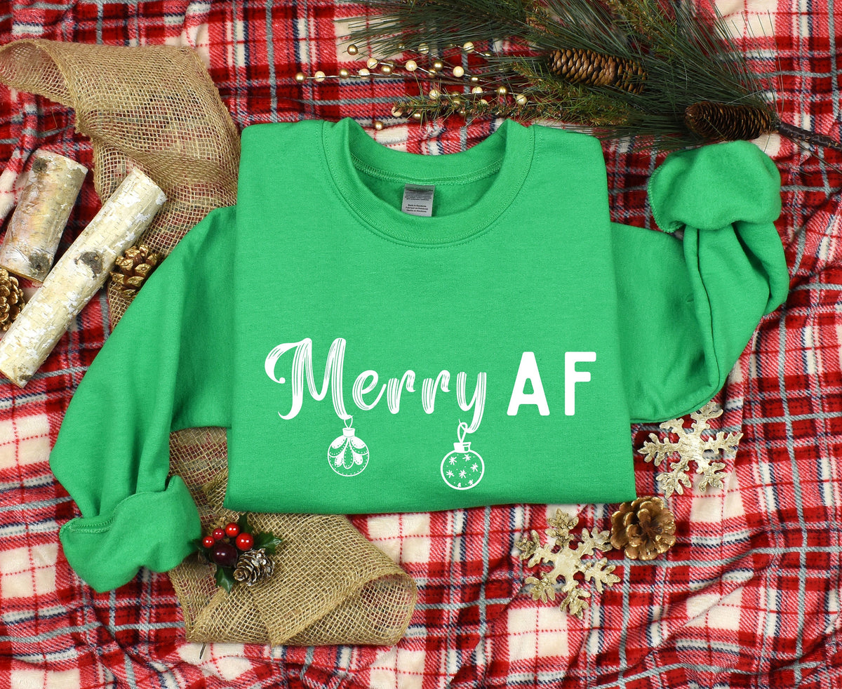 Merry AF Christmas Sweatshirt in Irish green laid flat on red plaid with pinecones, burlap, and gold snowflakes.