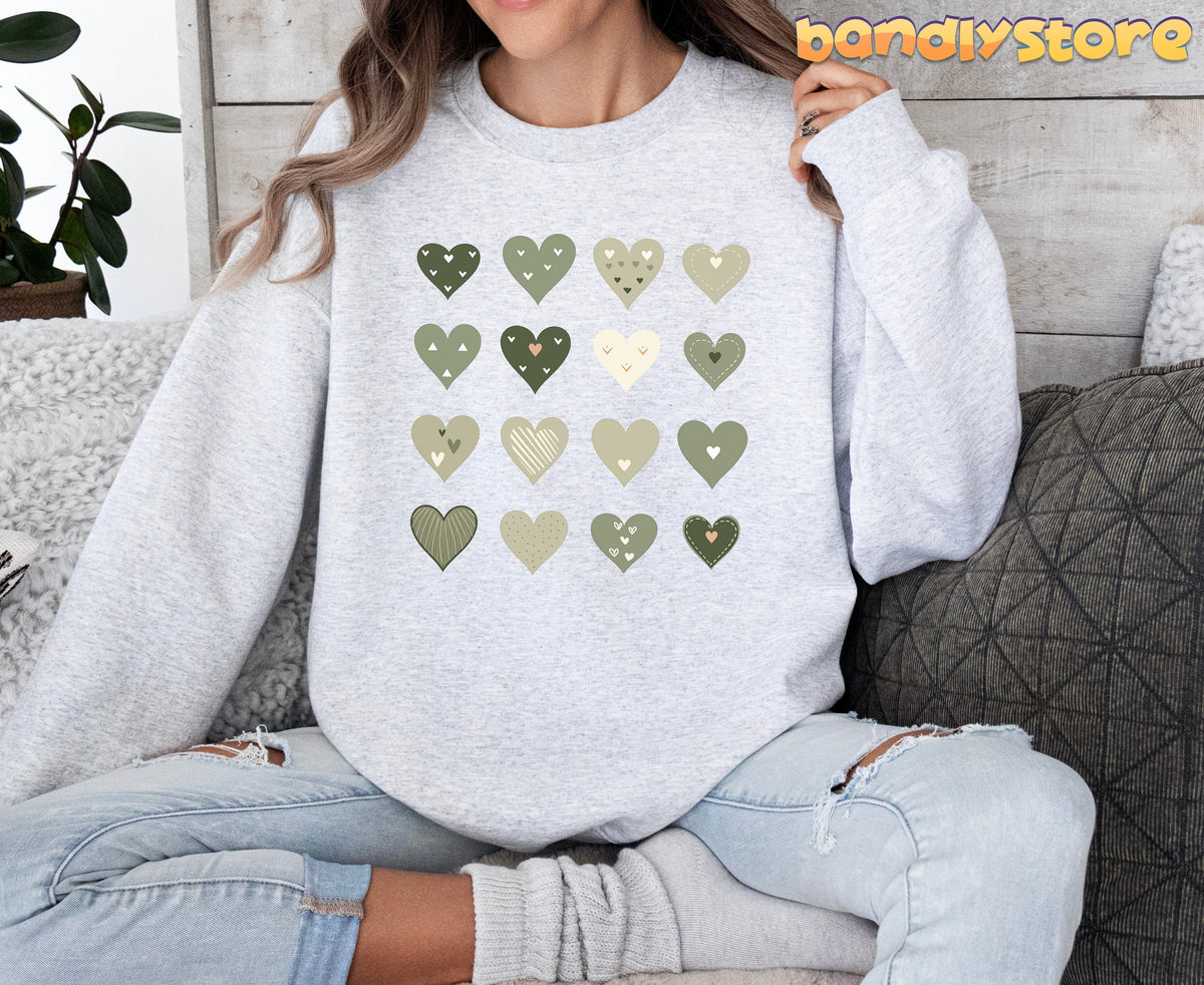 Minimalist Cute Hearts Sweatshirt in Sage Green worn in ash by a seated person on a couch with pillows and plant.
