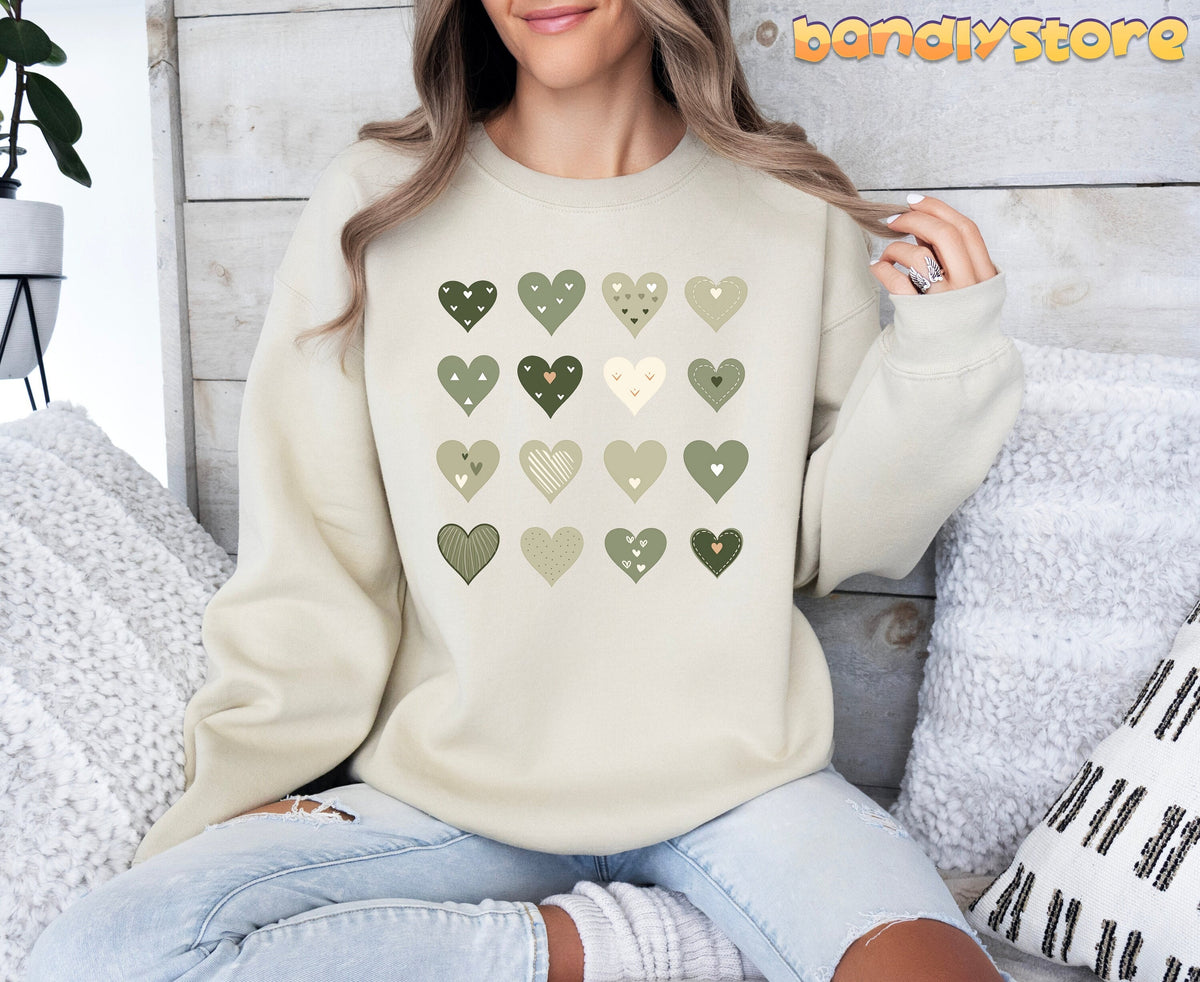 Minimalist Cute Hearts Sweatshirt in Sage Green on sand, front view of woman seated amid pillows by wooden wall and plant.